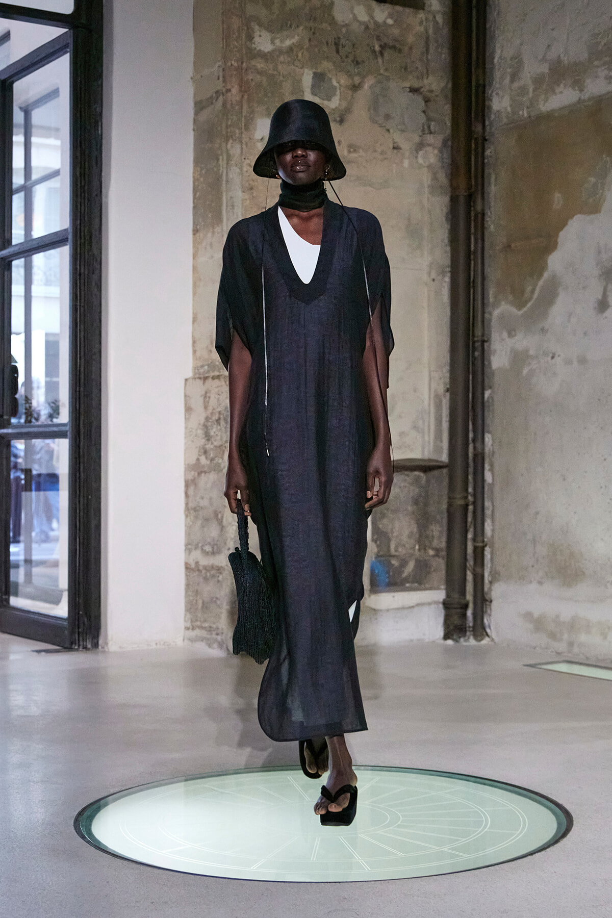 Model in a dark loose maxi dress over a white top, wearing a black bucket hat and carrying a black bag, walking on a lit circular platform.