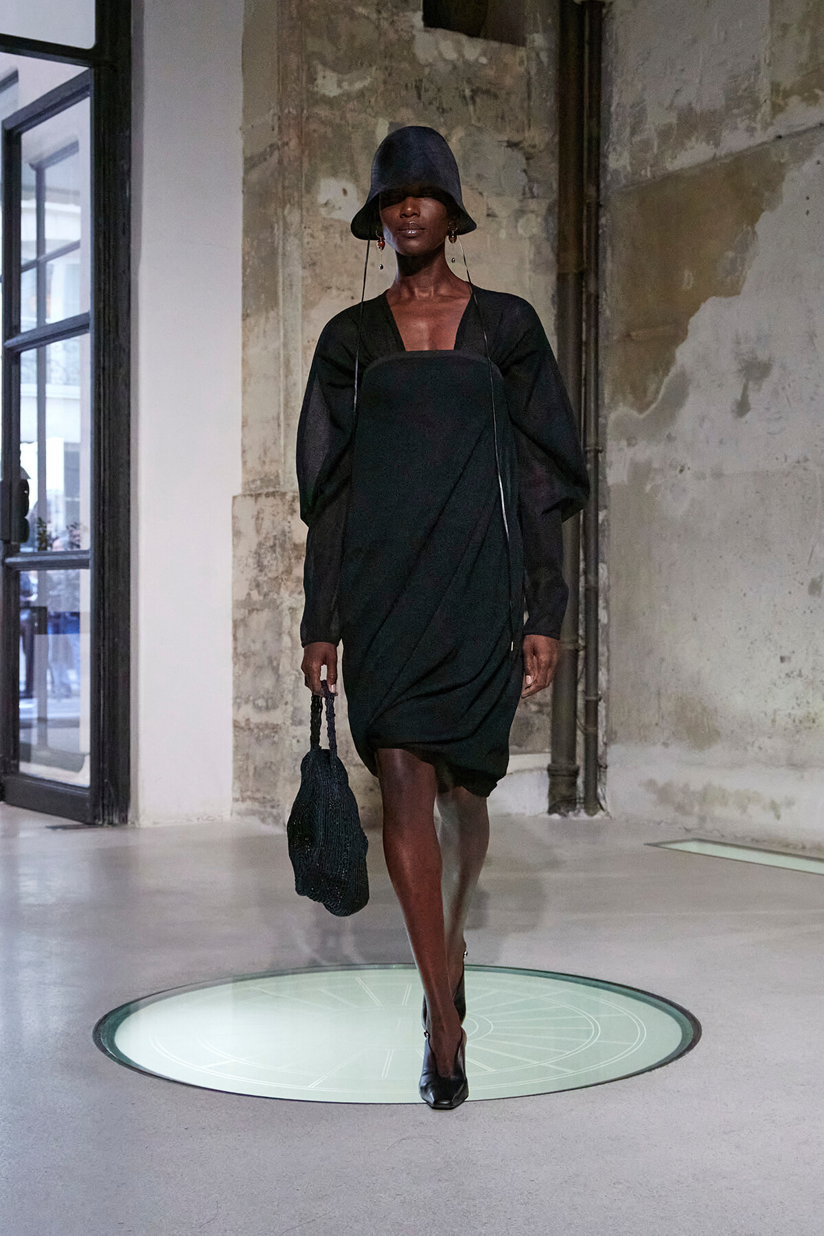 Model in a black draped dress and wide-brim hat walks a fashion runway in an industrial interior with exposed walls.