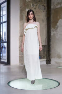 Model wearing a white off-the-shoulder pleated maxi dress, standing on a circular display platform in an industrial-style studio.