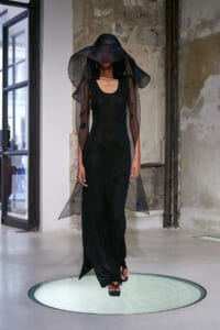 Model walks a black floor-length gown with sheer long sleeves and an oversized sheer veil hat on a minimalist runway in an industrial space