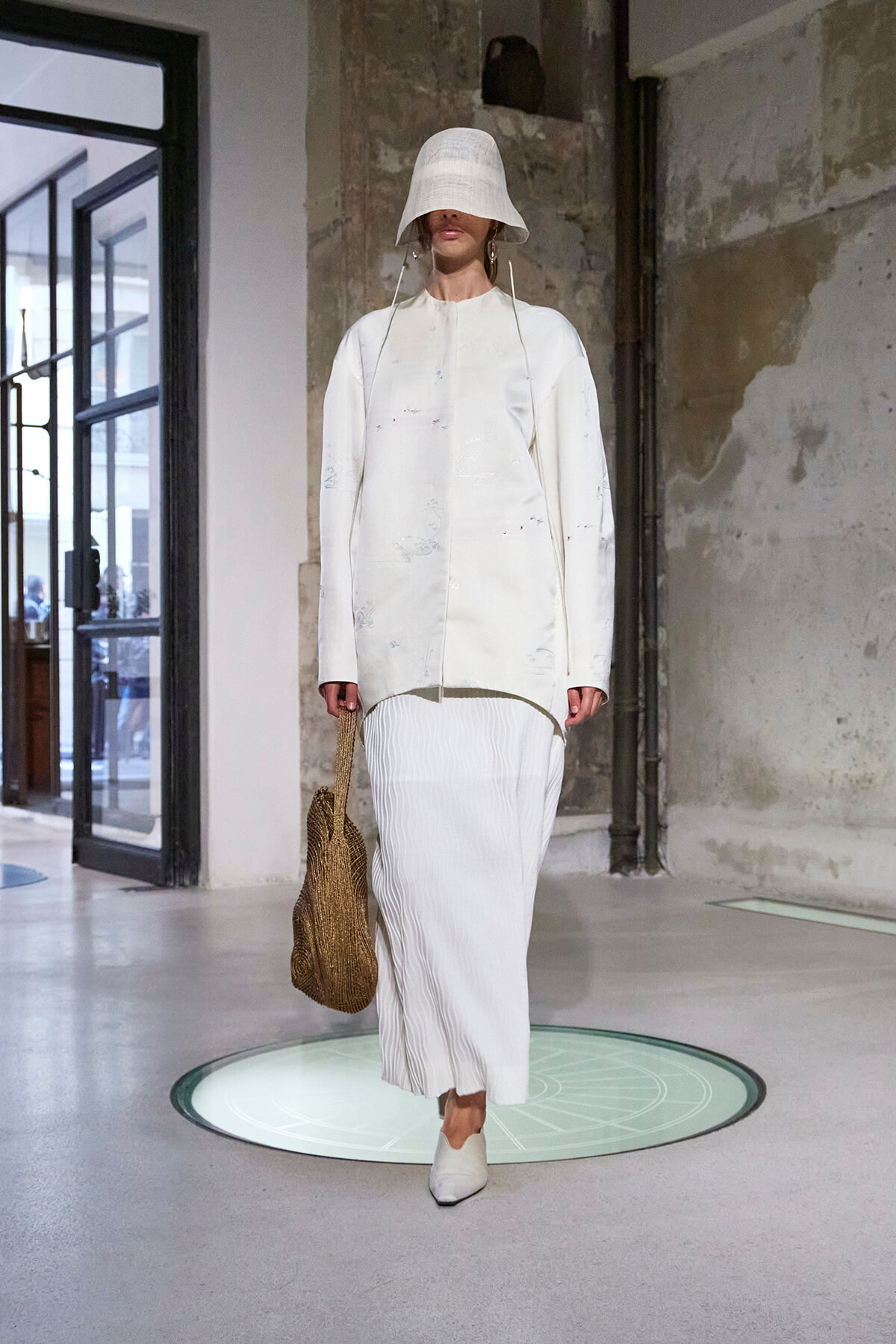 Model in an all-white ensemble: oversized coat with subtle paint specks, pleated midi skirt, and a white bucket hat, carrying a woven brown bag.