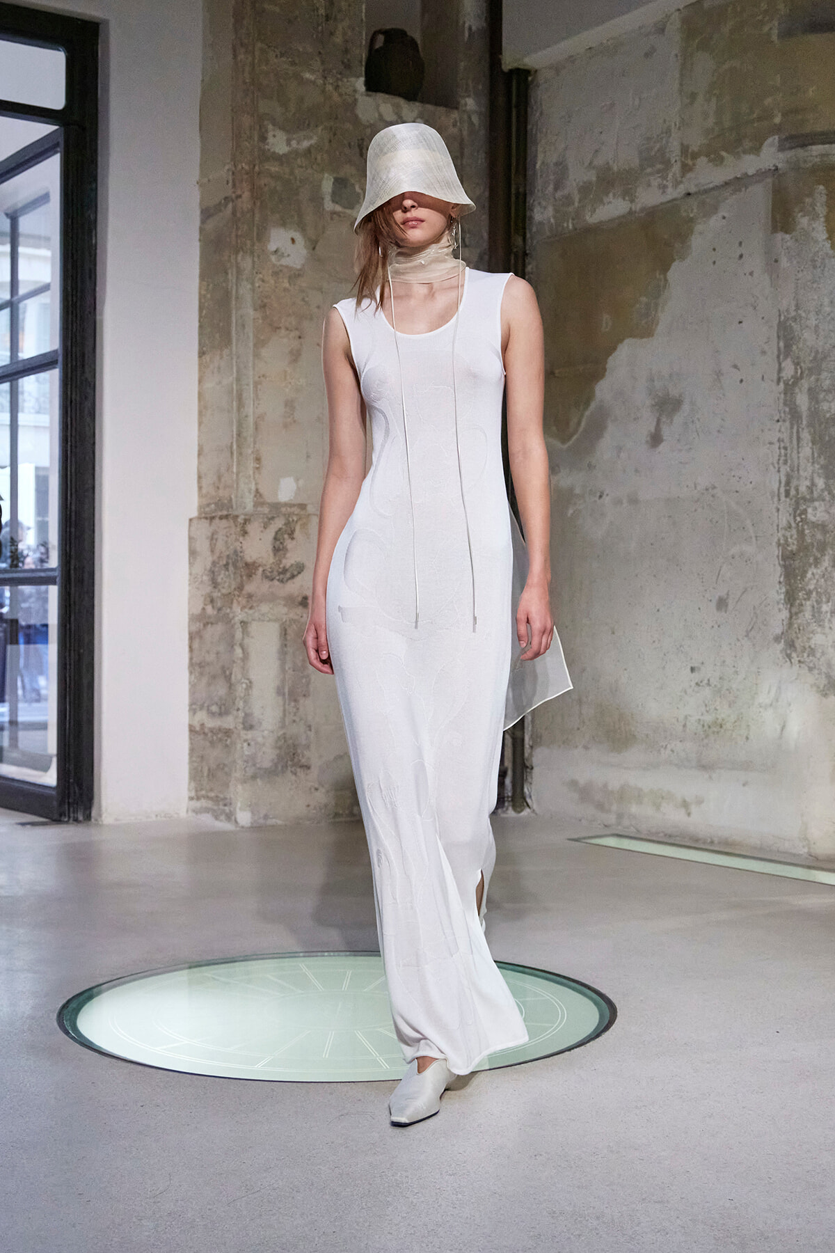 Model on a runway in an industrial space, wearing a long white sleeveless dress with subtle embroidery, a wide white bucket hat partially covering her face, and matching white shoes.
