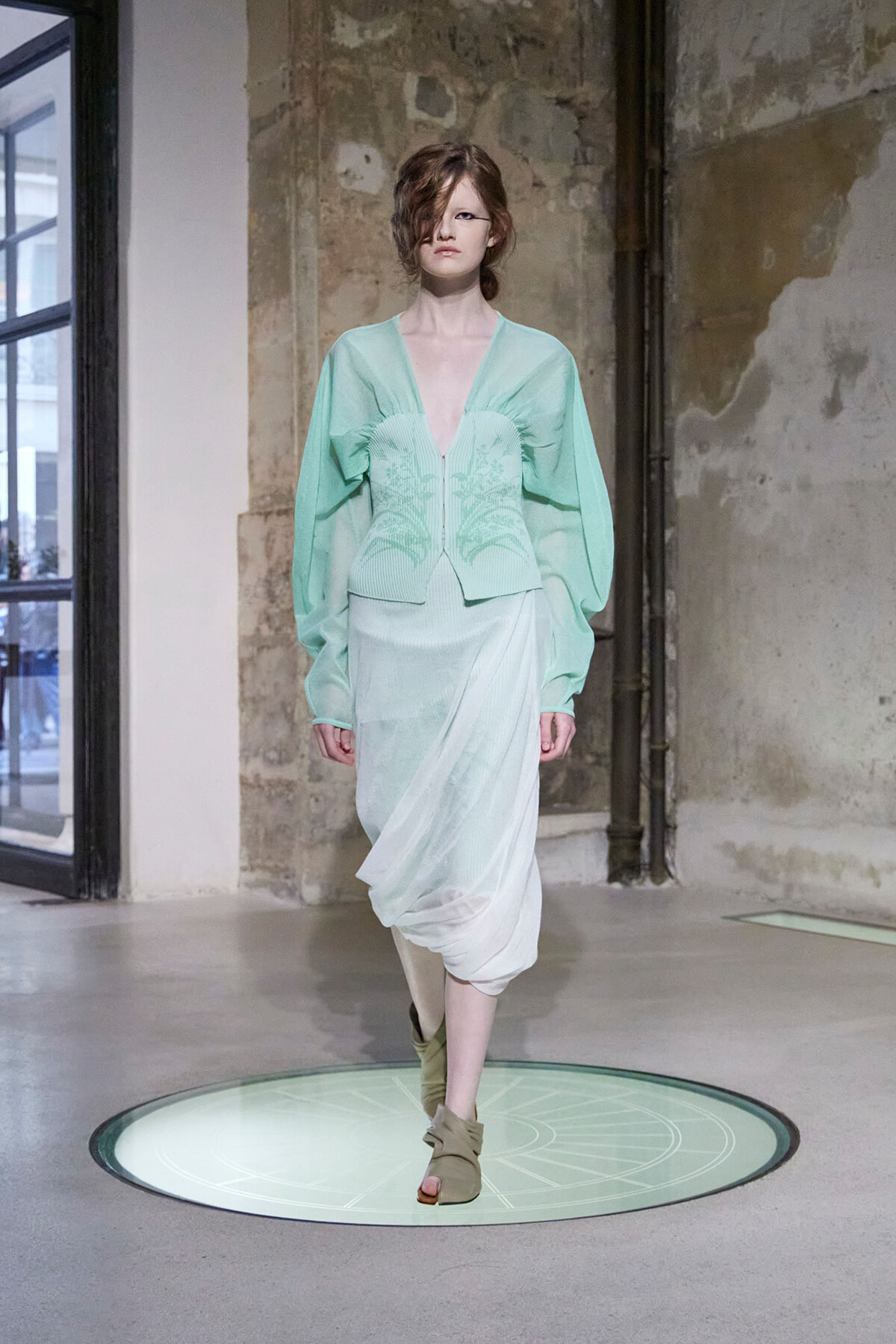 Model on a fashion runway wearing a mint-green, deep-V top with voluminous sleeves and a sheer draped skirt.
