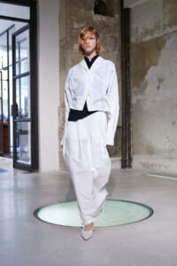 Model walking in a white, oversized blazer and loose white pants on a runway in an industrial space with exposed walls.