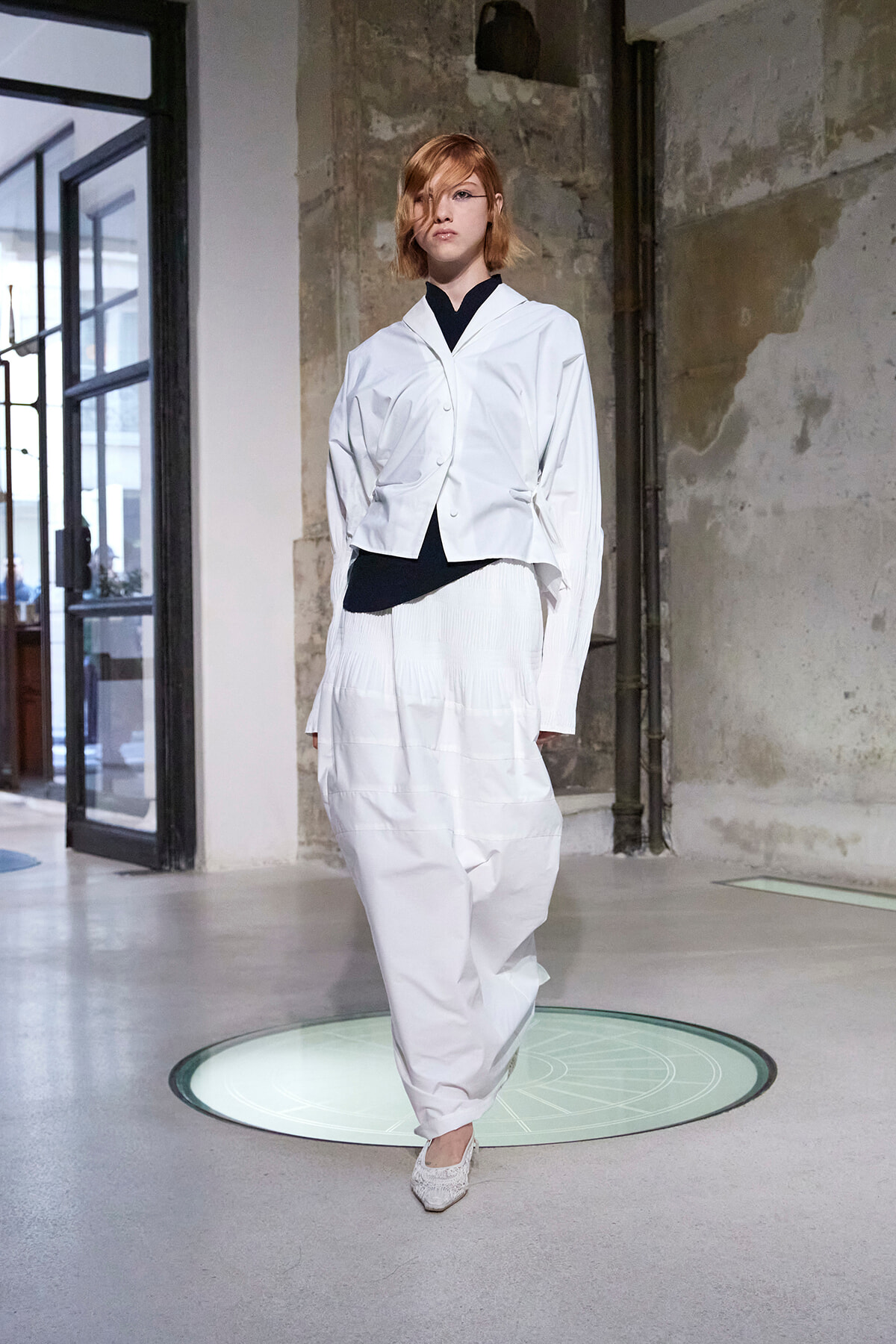 Model walking in a white, oversized blazer and loose white pants on a runway in an industrial space with exposed walls.