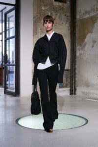 Model walks a runway in an industrial space wearing a black oversized jacket over a white top and black pants, carrying a black net bag.