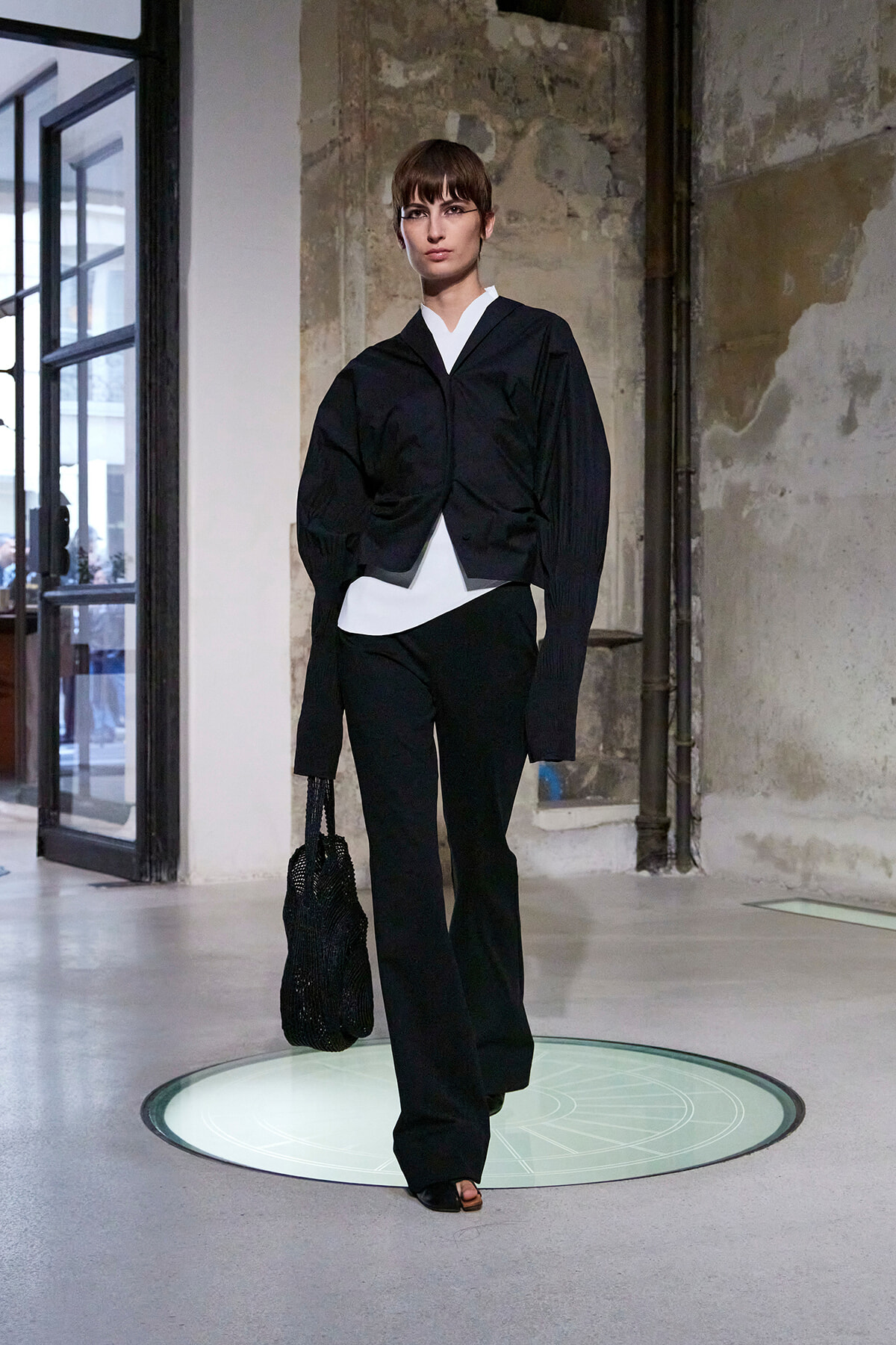 Model walks a runway in an industrial space wearing a black oversized jacket over a white top and black pants, carrying a black net bag.