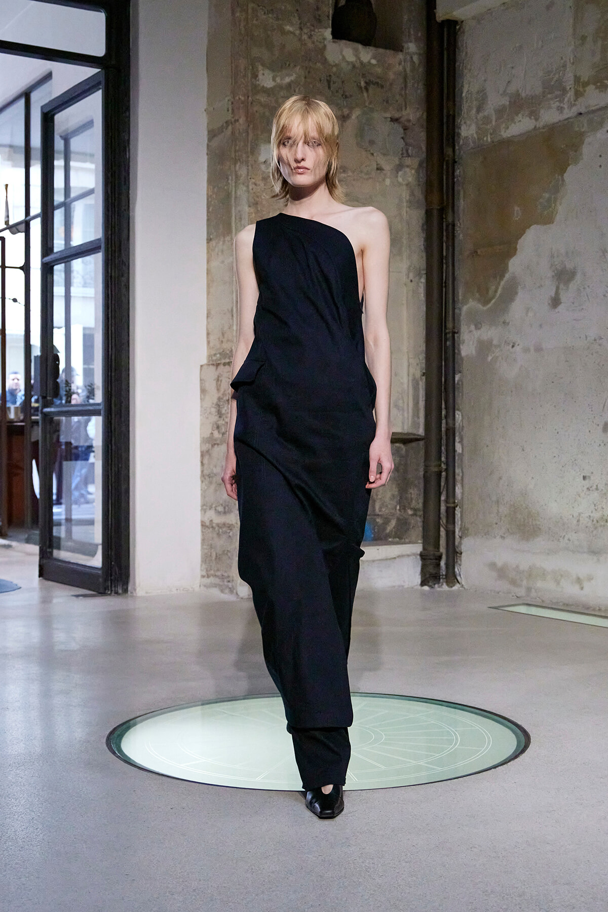 Model walking a black one-shoulder dress down a runway in an industrial, concrete space with exposed walls.