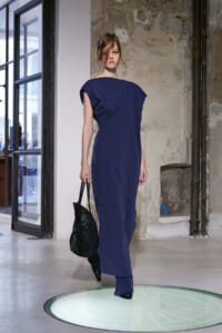Female model in a navy blue, one-shoulder sheath dress walks on a circular glass platform in an industrial loft, carrying a black woven handbag.