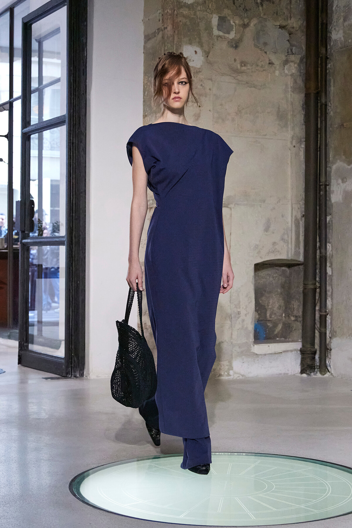 Female model in a navy blue, one-shoulder sheath dress walks on a circular glass platform in an industrial loft, carrying a black woven handbag.
