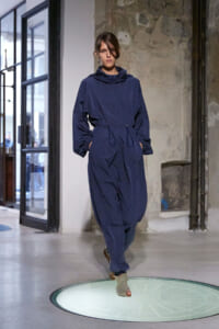 Model walks the runway in a navy blue hooded jumpsuit with a drawstring waist, industrial backdrop behind.