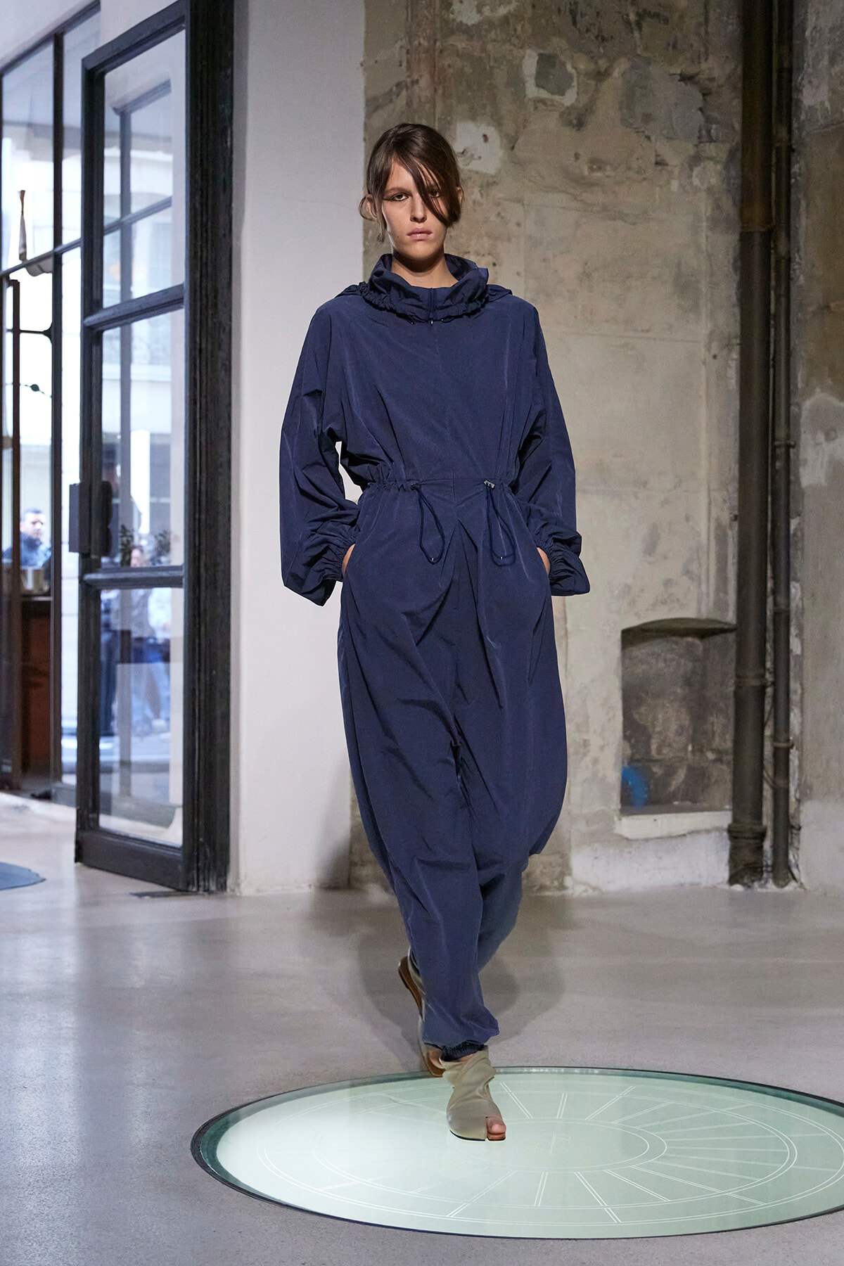 Model walks the runway in a navy blue hooded jumpsuit with a drawstring waist, industrial backdrop behind.