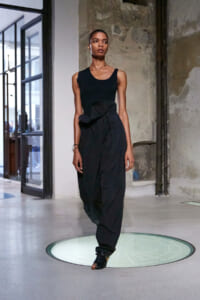 Female model walks the runway in a black sleeveless top and oversized black pants with a tied waist belt, in an industrial studio setting.