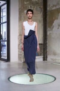 Model on a circular illuminated runway platform wearing a white sleeveless top and navy wrap skirt with a large bow. An industrial background is visible behind her.