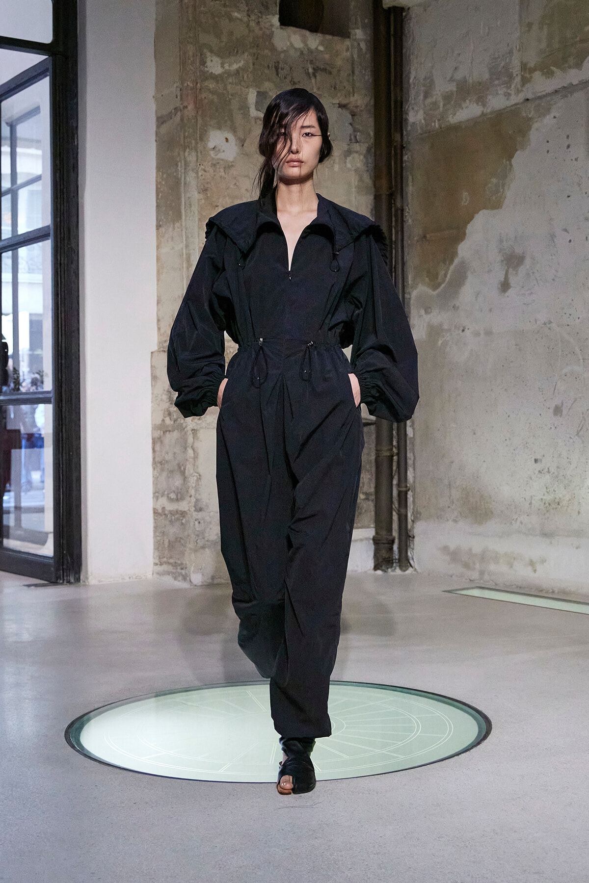 Model on a runway wearing a black jumpsuit with a cinched waist and voluminous sleeves in an industrial setting.