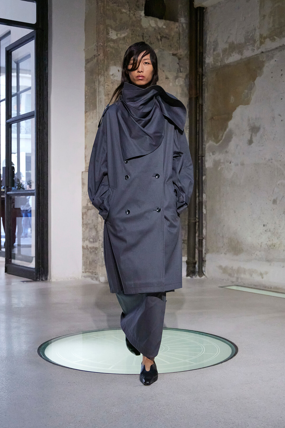 Model wearing a loose, double-breasted gray trench coat with a draped, high collar in an industrial interior setting.