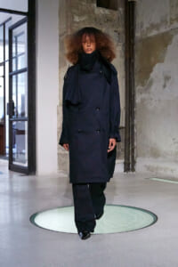 Model wearing a dark navy double-breasted trench coat with a black scarf, walking on a circular glass floor in an industrial studio.
