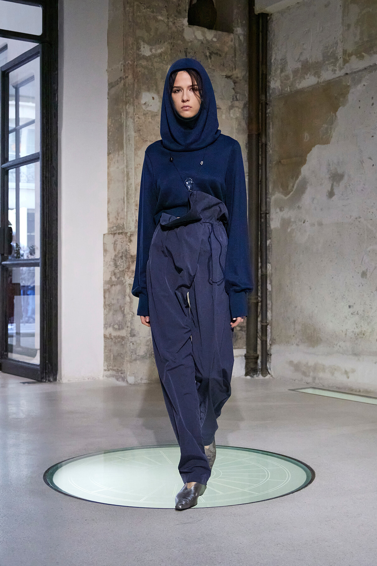 Model in a navy hooded top and loose navy pants walks a runway on a circular glass light platform in an industrial space.