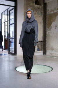 Model walks a runway in a gray hooded top layered over a black, wrap-style skirt in an industrial gallery setting
