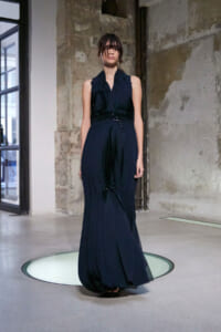 Model in a navy pleated evening gown with a beaded waist, standing on a circular glass platform in an industrial space