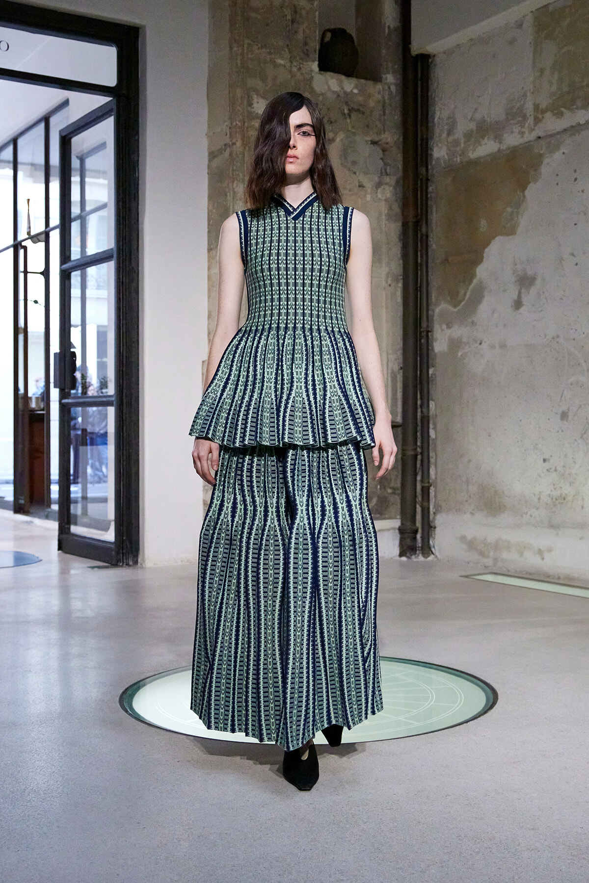 Model in a teal and navy sleeveless peplum top with a matching long skirt, standing on a circular glass platform in an industrial studio.