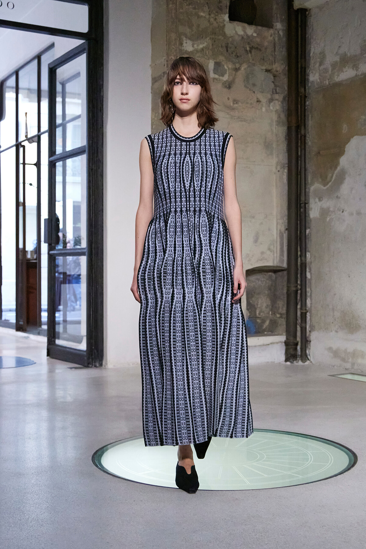 Model wearing a sleeveless black-and-white patterned maxi dress standing on a circular glass platform in an industrial setting