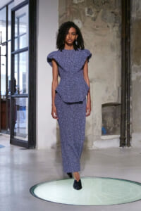 Model walking a blue patterned dress with oversized flutter sleeves on a circular runway platform in an industrial studio.