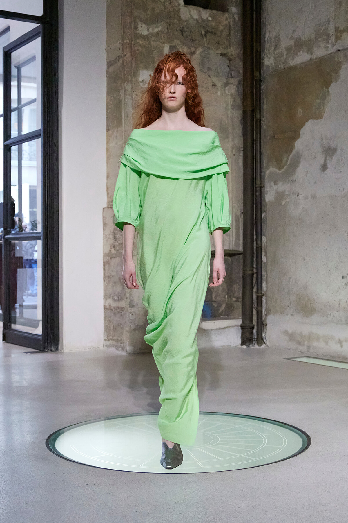 Model walks a runway in a lime-green off-shoulder draped dress with puff sleeves against an exposed brick wall backdrop.