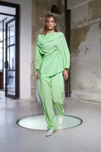 Model wearing a lime-green draped jumpsuit with an off-shoulder top and wide pants, white boots, and a silver mesh bag, walking on a circular lit platform in an industrial studio.
