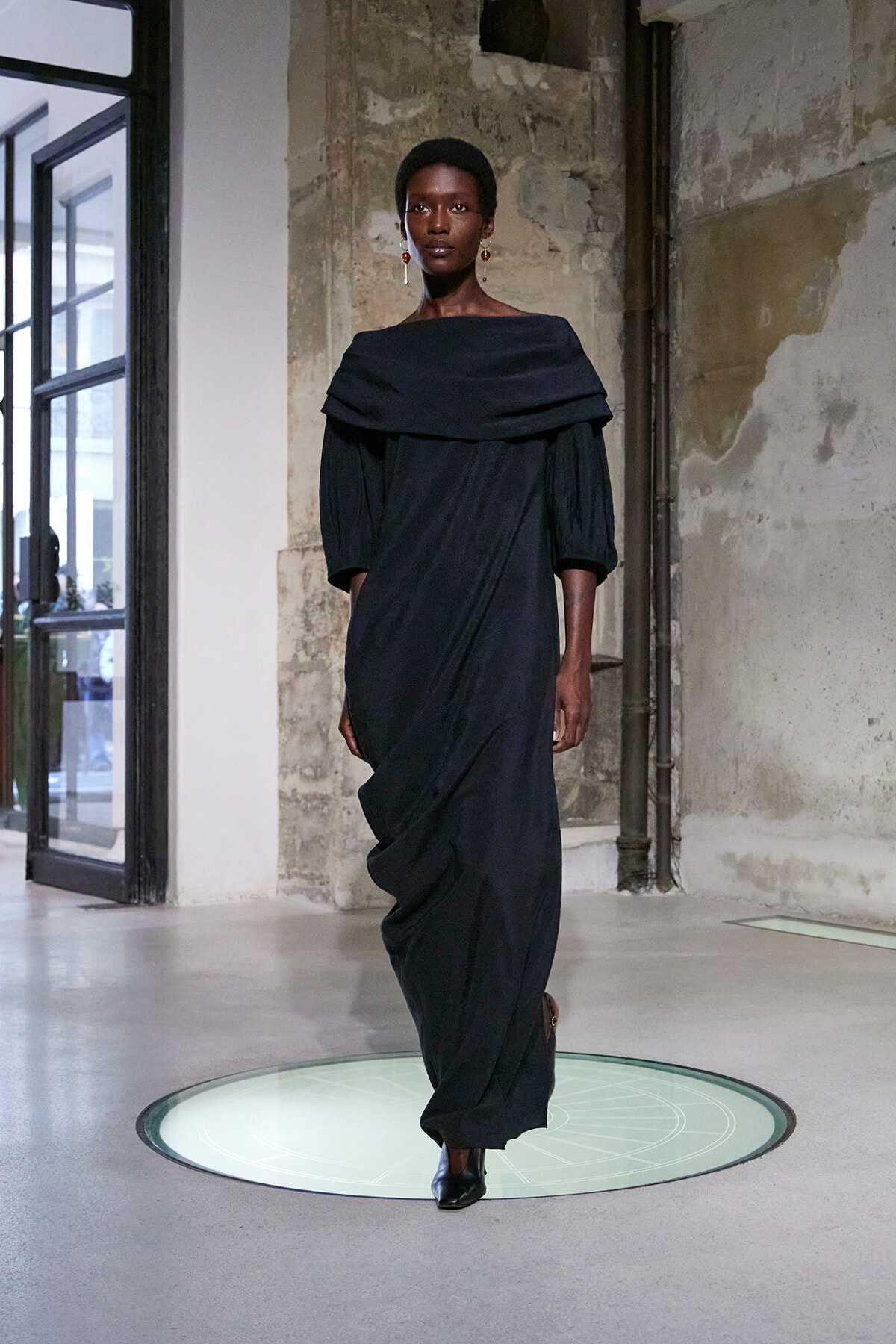Model in a dark off-shoulder draped gown walks on a glass-lit runway circle in an industrial studio setting.