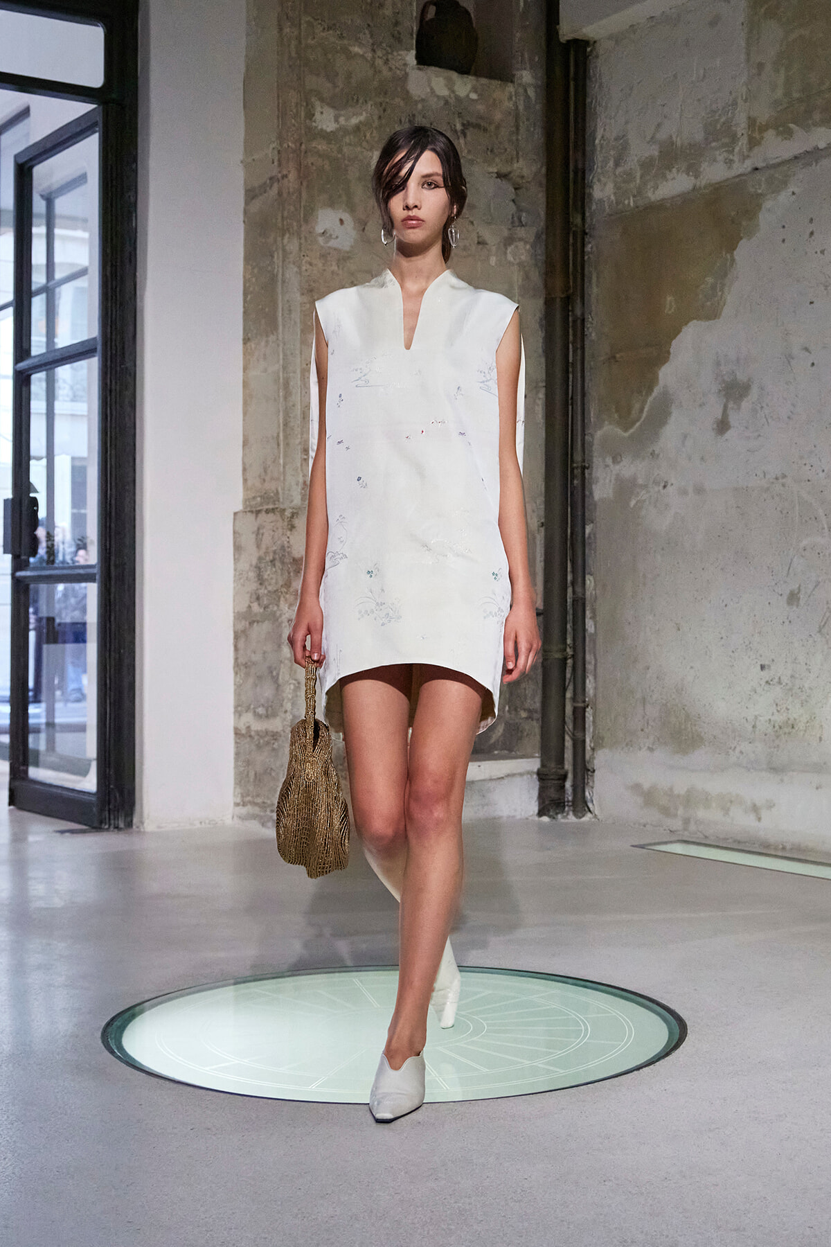 Model walks the runway in a white sleeveless shift dress with subtle embroidery, carrying a woven handbag, in an industrial venue.