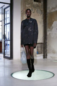 Model walks down a runway in an oversized black tunic with subtle white floral specks, paired with black shorts and knee-high boots, carrying a gray mesh tote bag.