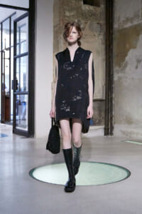 Model walking a runway in a sleeveless black dress with subtle white embroidery, knee-high boots, and a black bag in a gritty venue.