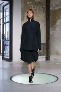 Model in an oversized dark tunic with a high collar walking on a circular illuminated runway in an industrial space.