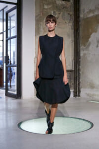 Model walks on a circular glass runway in an industrial studio, wearing a dark sleeveless dress with a layered peplum and ankle boots.
