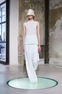 Model in all-white sleeveless top and wide-leg pants, wearing a bucket hat, walking on a fashion runway in an industrial setting.