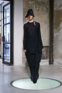 Model walking a runway in an all-black, oversized outfit with a wide-brimmed hat in an industrial space on a round, lit platform.