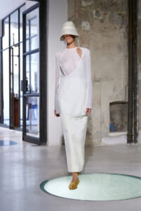 Model walking a runway in an all-white outfit: sheer long-sleeve top with a wrap-front over a white dress, bucket hat covering eyes, and light-colored skirt in an industrial space.