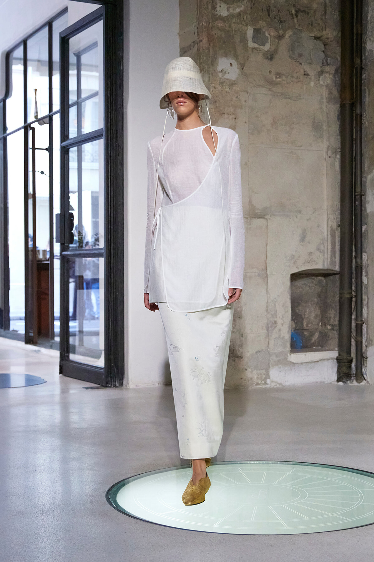 Model walking a runway in an all-white outfit: sheer long-sleeve top with a wrap-front over a white dress, bucket hat covering eyes, and light-colored skirt in an industrial space.