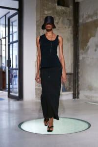 Model walks a circular glass runway in an all-black look: sleeveless maxi dress, oversized bucket hat, in an industrial studio.