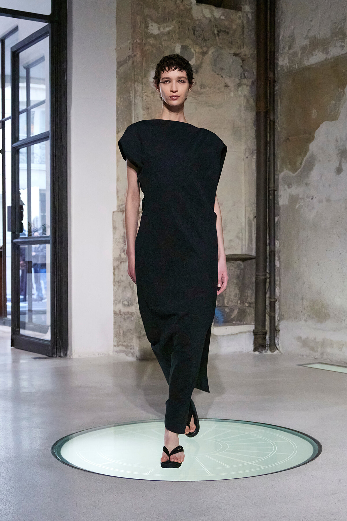 Model walks a minimalist black dress down a runway in an industrial space, standing on a circular glass floor panel.