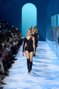 Model in a black asymmetrical bodysuit and knee-high boots walks a blue-lit runway, audience on both sides filming with phones.