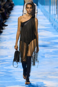 Model walks the runway in a black one-shoulder fringe dress with long beaded chains, paired with knee-high boots and a small black fringe bag.