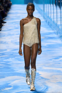 Model walking the runway in a metallic gold one-shoulder fringe bodysuit with tall lace-up boots on a blue-patterned catwalk.