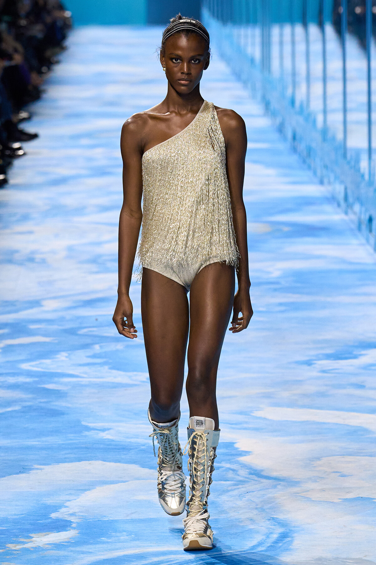 Model walking the runway in a metallic gold one-shoulder fringe bodysuit with tall lace-up boots on a blue-patterned catwalk.