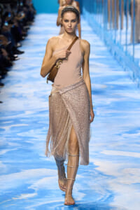 Model walks the runway in a pale pink one-shoulder top, a sparkling wrap fringe skirt, and strappy sandals, carrying a brown crossbody bag.