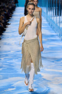 Model on a fashion runway wearing a white tank top, silver fringe skirt, white knee-high boots, and a white shoulder bag.