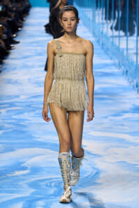 Model walking the runway in a short, gold fringe strapless romper with a beige one-shoulder strap, paired with tall, light-colored lace-up boots on a blue-toned catwalk.