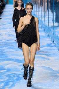 Model walks the runway in a black one-shoulder fringe bodysuit, paired with black lace-up boots and a shoulder bag against a blue-toned runway backdrop.