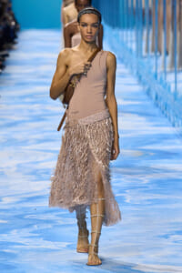 Runway model walks the catwalk in a beige one-shoulder top and a pale pink fringe skirt, carrying a brown crossbody bag.